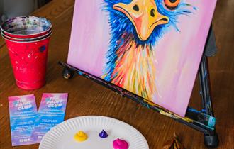 Painting class set up on a table including a tabletop easel with a finished landscape painting on canvas with red cup with paintbrushes neatly scatter