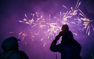 A family watching low bangs fireworks at Godstone Farm. The sky is purple and there are pretty fireworks. There is a lady and a man who has a child on