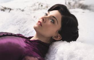 A young woman lies in the snow, looking up to the sky. She wears a rich purple silk dress with a high collar. She has brown hair and striking blue eye