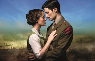 A couple embrace lovingly with solemn facial expressions. The man on the right wears military uniform and has dark hair and a moustache. The woman on