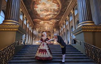 A man dressed in 18th-century attire is bowing to a woman in a lavish red and gold period gown on an ornate staircase in the Painted Hall.