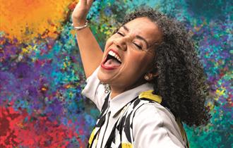 A woman sings joyfully, eyes closed and mouth open wide. Her head is passionately thrown back and her arm is raised. She has black and grey curly hair