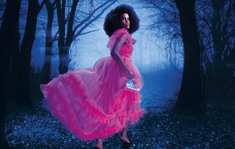A woman in along, ruffled pink chiffon dress runs through a dark, misty woodland away from the camera. She is turning to look behind her, has an afro