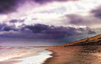 A moody watercolour image of a coastline under stormy skies. The sandy cove and dunes curve away into the distance, with waves breaking gently on the