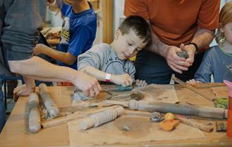 Young boy sitting at a table, stamping a pattern into a slab of clay. His parents are standing either side of him.