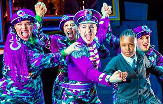 Four women in purple combat style trousers and purple military style jackets and hats are semi-crouched, facing out front, and clutching the hand of a