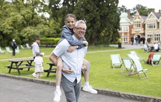 A father carries his daughter on his back as they walk past the Mansion at Bletchley Park. Both are smiling towards the camera.