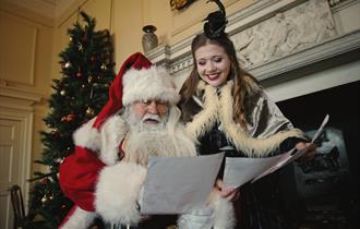 A man dressed as Father Christmas, with a traditional red and white Santa suit and a long white beard, is sitting by a fireplace. He is reading letter