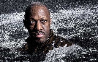 A man's head and shoulders rise from a sea of rough, churning water; rain is pouring down on him. He stares into the distance, his face illuminated by