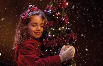 A young girl in a red winter coat hugs a small Christmas tree. She is smiling contentedly with her eyes closed, and has a red tartan bow in her hair.
