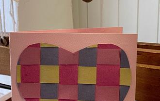 image of heart woven from paper on a card which is sitting on top of a wooden weaving loom