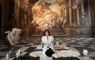 A person dressed in a white robe sits cross-legged on a white fur rug in an ornate hall with checkered floors. A large classical mural decorates the w