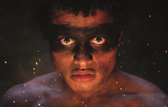A boy stares brutally straight into the camera with a straight face. There is messy black dirt over his eyes, like a mask, and he is topless with dirt