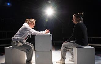 Two women sit on white blocks on a bright white stage; the background is in darkness but there's a central stage light glaring out front. The woman on