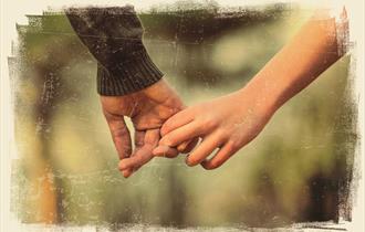 Two hands with two fingers linking, pulling away slightly. One has a bare arm and the other wears a khaki jumper. The background is dappled green and