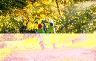 Two children run through autumnal woodland at Wakehurst wearing Nature Heroes capes