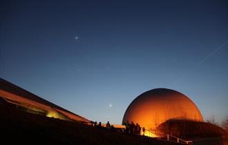 A dark sky with a few stars. The Science Centre and Planetarium are glowing orange against the night sky.