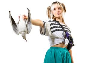 A young woman with blond hair tied up in bunches stares straight out front with a seagull thrust out in front of her, held in her right hand. She wear