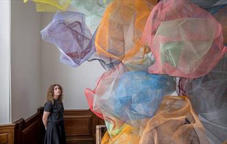 A women looks up a structure made from coloured mesh clouds that seem to float in the middle the stairwell of a wooden staircase.