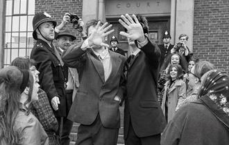 A black-and-white-photo taken on the crowded steps of Chichester Law Court. In the centre, two young men in stylish suits walk down the steps, holding