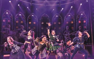 Six women in modern versions of Tudor outfits pose like a pop group, singing into handheld microphones and dancing in a variety of lively and energeti