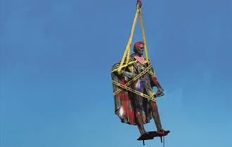 A traditional stone statue of a historic figure is suspended from an orange metal hook with yellow straps against a bright blue background. It is wrap