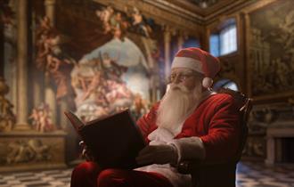 Father Christmas reading a book inside the Painted Hall. He looks intrigued and engaged.