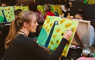 Person smiling, relaxed, sitting down in a group setting with a canvas on an easel with paintbrushes in a c up beside it