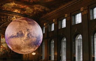 In a grand, ornate hall with a high, frescoed ceilings (the Painted Hall), a large, glowing model of planet Mars is suspended in the centre of the roo