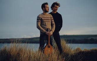 Two middle aged white men with dark hair and facial hair stand on the cliffs looking wistfully into the distance