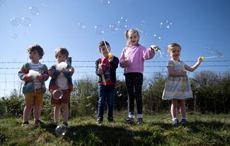 5 children stood outdoors with bubble wands and bubbles around them. The sky is bright blue in the background. 2 boys are holding foam in their hands.