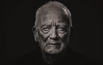 An elderly man (Ian McDiarmid) stares at the camera. His face is wrinkled and looks stern and dramatic, with shadows under his eyes and chin.  He is w