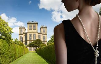 Blue sky with white clouds are the backdrop to a stateley home, with green manicured lawns stretching out in front, at the foreground of the image. To