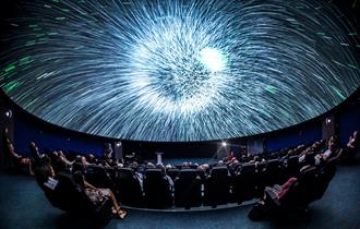 A blast of white light on a Planetarium dome. There are people sat in seats watching the screen.
