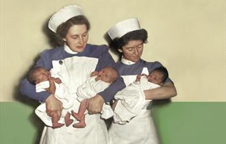 Two midwives in white smocks, white caps and blue blouses stand against a green and white wall. The midwife on the left holds two newborn babies, they
