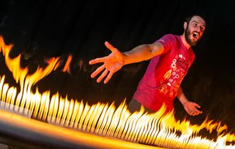 A man with a pink T-shirt on is standing in front of a large metal tube that has lots of holes in it. There is fire coming out of the holes and the ma