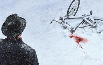 A black bicycle lies on the ground, half covered in snow. Next to the bike, a patch of snow is stained red with blood. In the foreground, a man is tur