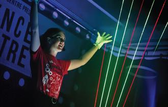 A science communicator stands with her arms in the air behind some coloured laser beams