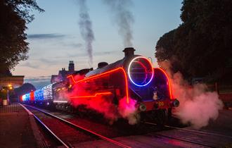 A steam locomotive travelling along a track with neon and fairy lights on the train.