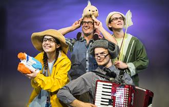 Four brightly dressed people, all wearing glasses, holding an array of objects: to the left, a smiley woman in a yellow raincoat and rain hat holds an