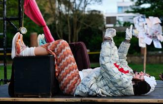 A person dressed in colourful clothing reclines on their back with legs propped up on a black box, pointing upward. The background includes trees, a b