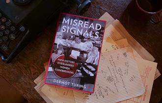 A book called 'Misread Signals' by Dermot Turing resting on a wooden desk with various bits of paper with codebreaking on