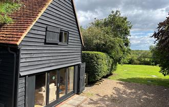 Black cladded barn with wide terrace doors. The view is a green field with trees in the far distance.