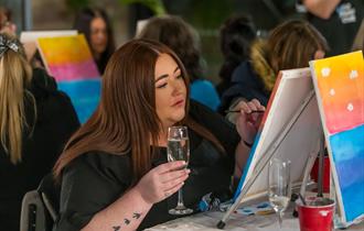 Person smiling, relaxed, sipping a drink sitting down in a group setting with a canvas on an easel with paintbrushes in a c up beside it