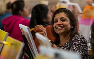 Person smiling, relaxed, sitting down, painting in a group setting with a canvas on an easel with paintbrushes