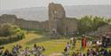 Crowd gathered on grass with stage and performers in front of Hastings Castle ruins in warm evening light.