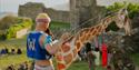 Performer holding a giraffe puppet and microphone at an outdoor event beside Hastings Castle ruins.