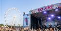 Crowds watching a band on the River stage with the wheel ride in the background at the Isle of Wight Festival, music event, what's on
