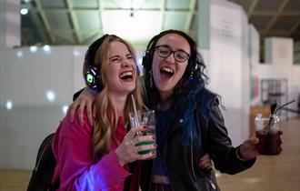 Two girls with headphones on singing and holding drinks