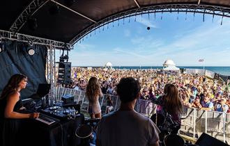 Photograph taken from within outdoor stage, look out at large crowd with the sea in the background.
